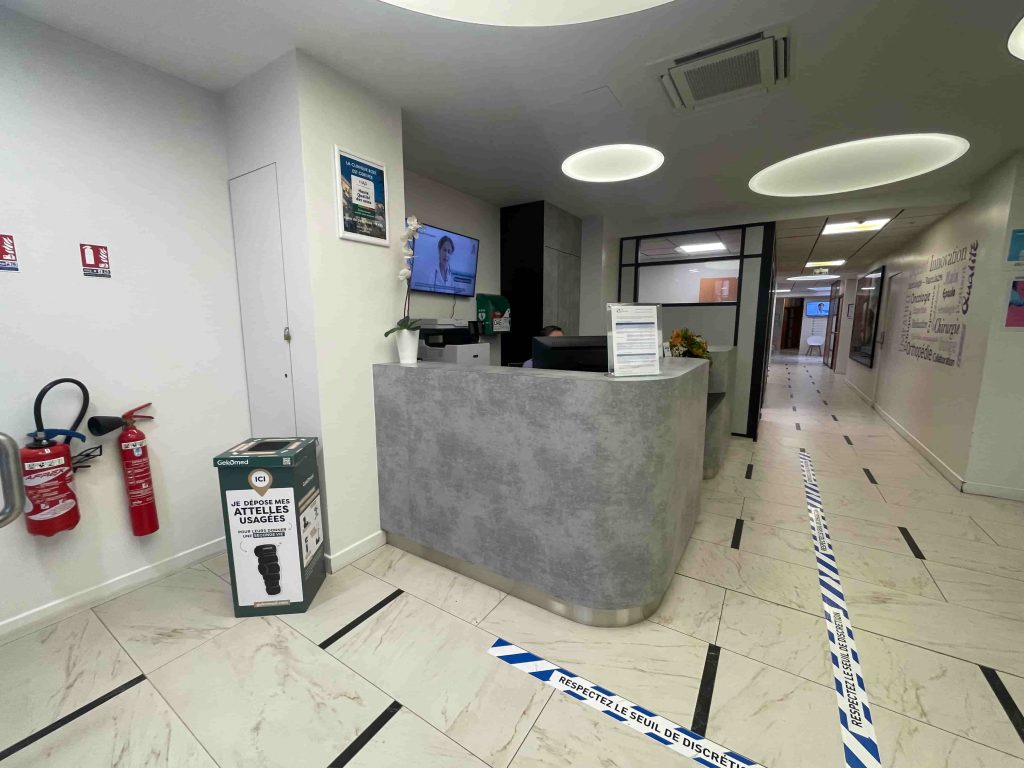 Borne de collecte Gekomed pour attelles usagées installée à l’accueil d’une clinique, à côté du comptoir de réception.